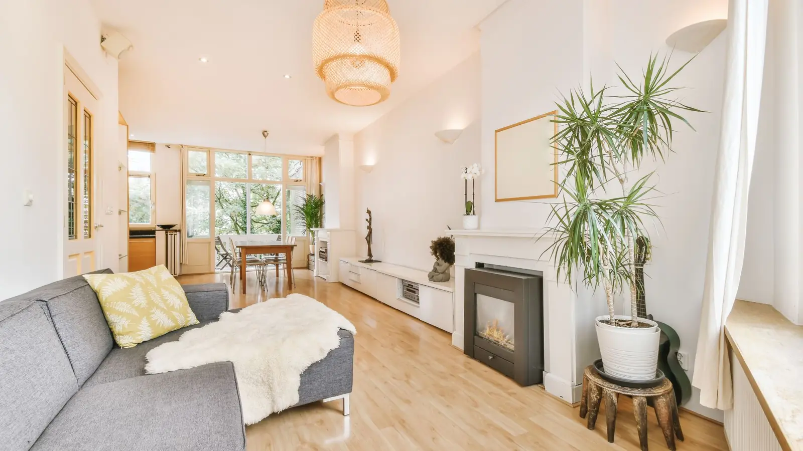 A sunlit living room with a fireplace, a gray sofa, tall plants and windows onto the trees.